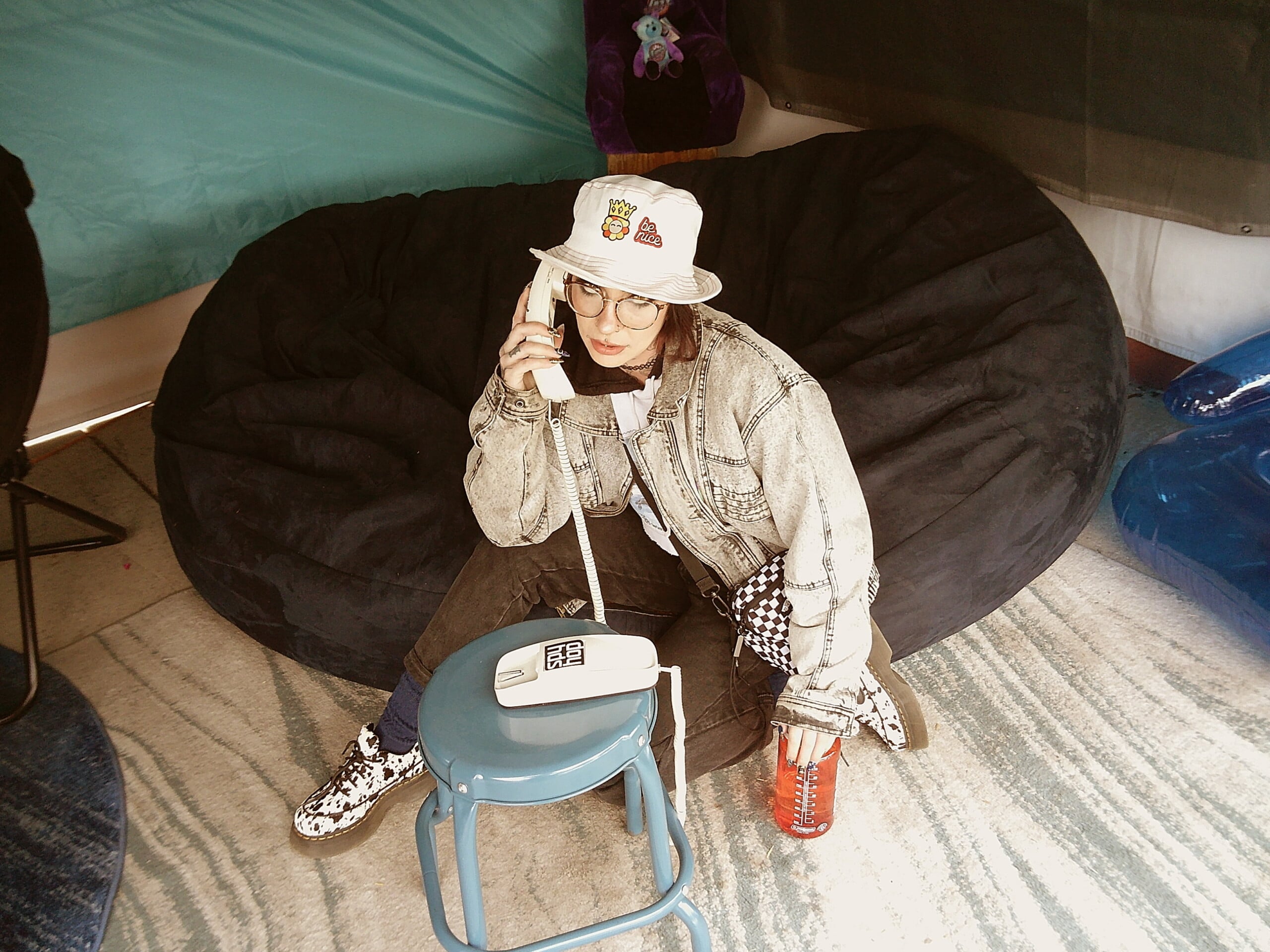 A person wearing a jean jacket, patterned sneakers, and a bucket hat sits on a black beanbag, holding a vintage landline phone and a red drink, with a blue stool and a soft, patterned rug nearby.