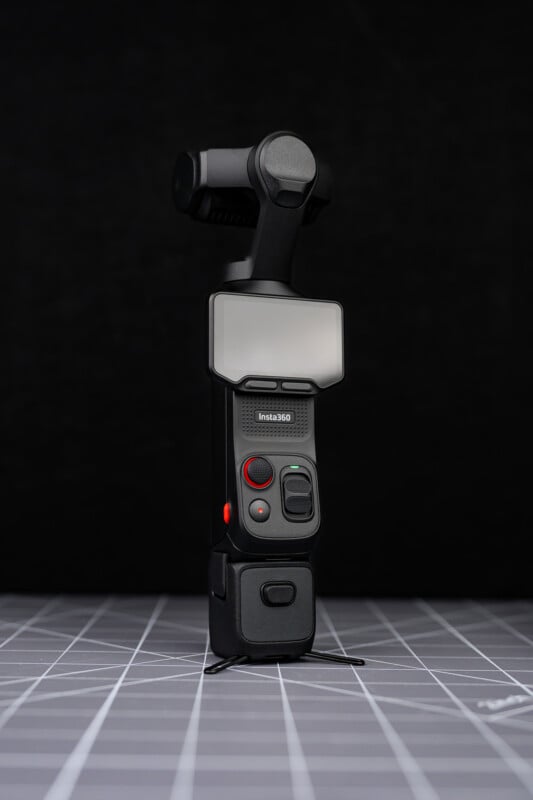 A compact black Insta360 camera with a gimbal and display screen stands upright on a grid-patterned gray surface against a dark background.