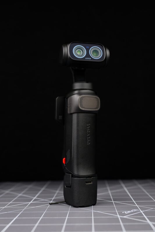 A compact black handheld camera with dual lenses on a small gimbal, standing upright on a gray grid-patterned surface against a dark background.