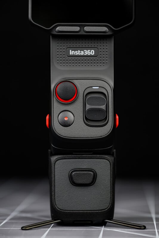 Close-up of an Insta360 camera on a small tripod, showing control buttons and textured surfaces. The camera is upright, with a dark background and a checkered surface below.