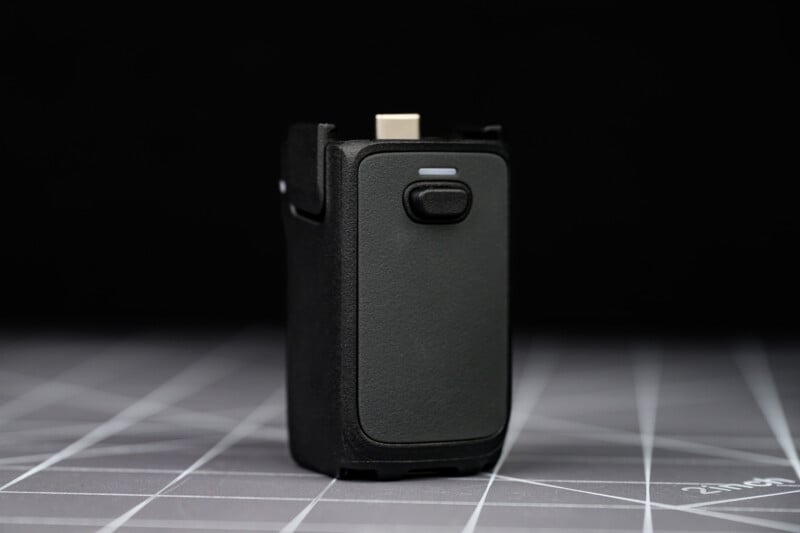 A close-up of a small, black electronic device with a single button on the front, standing upright on a gray, grid-patterned surface against a black background.