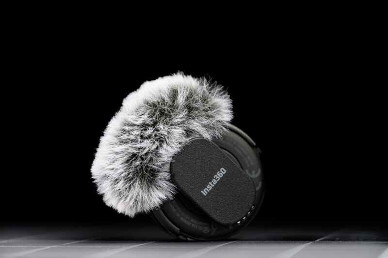 A close-up of an Insta360 microphone with a fluffy windscreen cover, set against a black background and resting on a reflective surface.