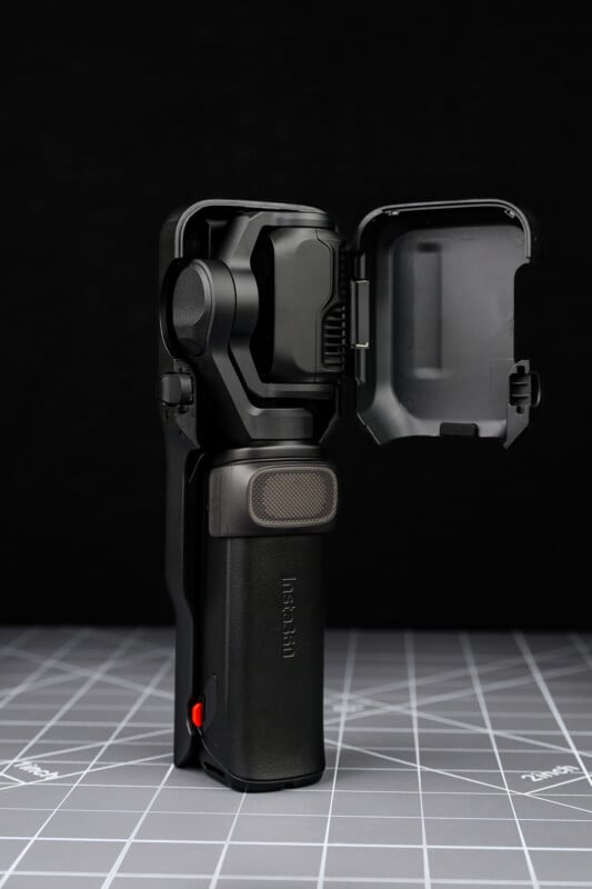 A black handheld gimbal camera with the protective cover open, standing upright on a gray grid-patterned surface, against a dark background.