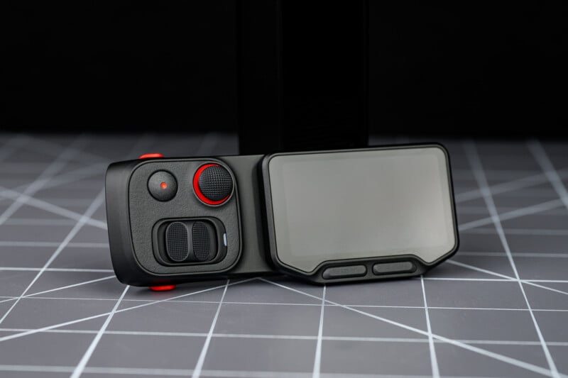 A close-up of a modern black electronic device with a large screen and red-accented buttons, placed on a gray grid-patterned surface against a dark background.
