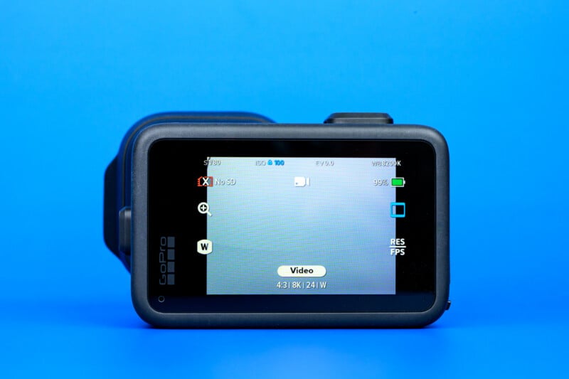 A GoPro camera with its rear touchscreen display facing forward, showing video recording settings on the screen, set against a solid bright blue background.