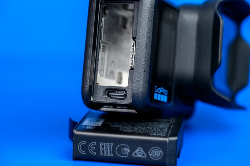 A close-up of a GoPro camera with its battery compartment open and the battery partially removed, against a blue background.