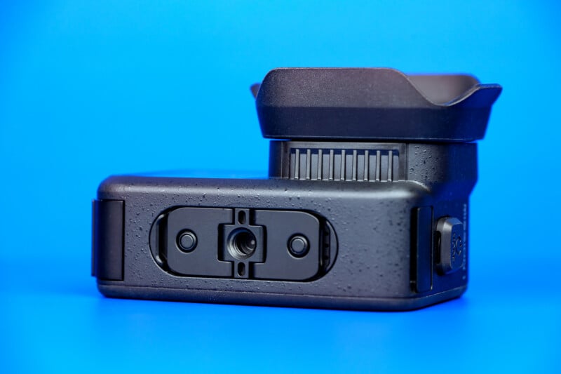 A close-up of a black camera or camera accessory, viewed from below, showing the tripod mount and textured surface, set against a bright blue background.