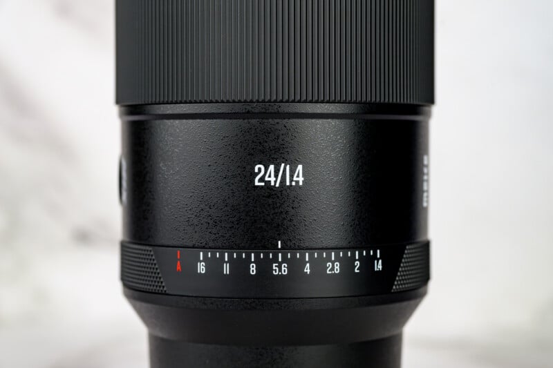 Close-up of a black camera lens showing "24/1.4" and an aperture scale ranging from f/1.4 to f/16, with textured and ribbed surface details visible.