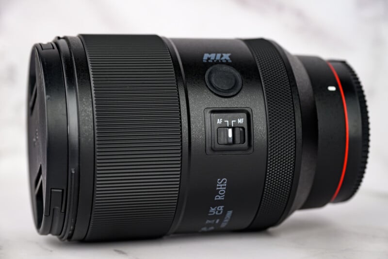 A close-up photo of a black camera lens with a textured zoom ring, AF/MF switch, and a red accent near the mount, placed on a white surface with a soft background.