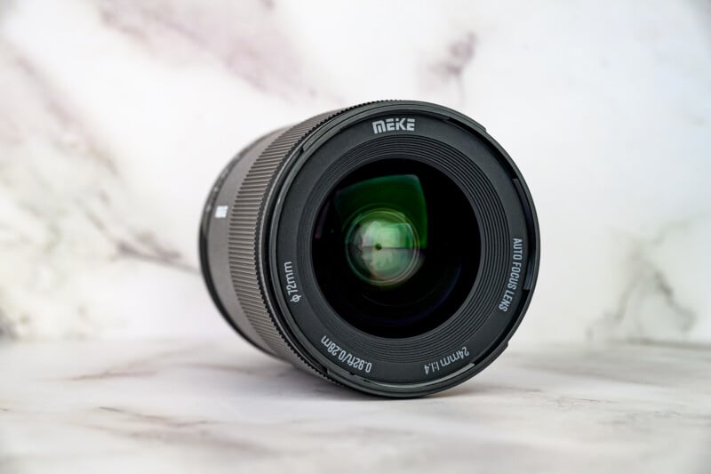 A close-up photo of a camera lens lying on a white marble surface. The lens front shows brand "MEIKE" and details like "85mm F1.4," "Ø72mm," and "DS-AF-85/1.4 STM." The background is softly blurred.