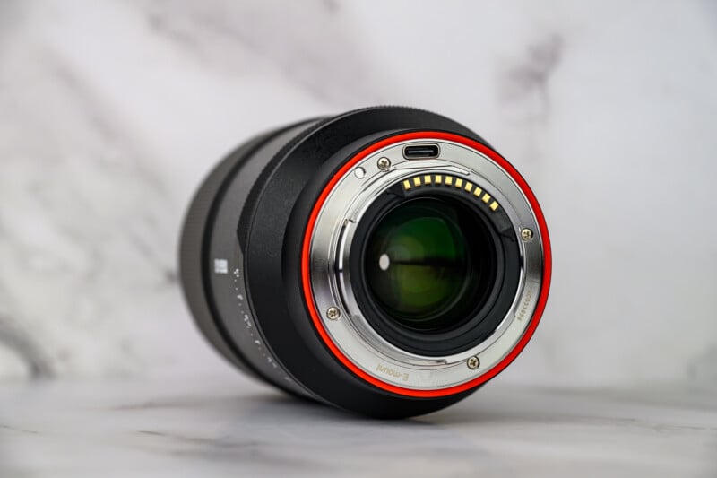 A close-up of a camera lens lying on its side, showing the electronic contacts and metal mount ring with a red accent; the background is soft and out of focus.