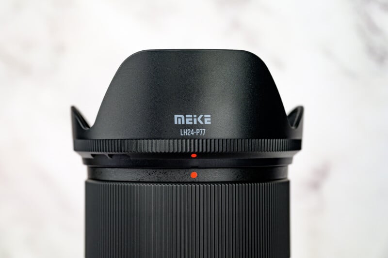 Close-up of a black Meike camera lens with a petal-shaped lens hood, labeled "MEIKE LH24-PT7," against a blurred, light-colored background.
