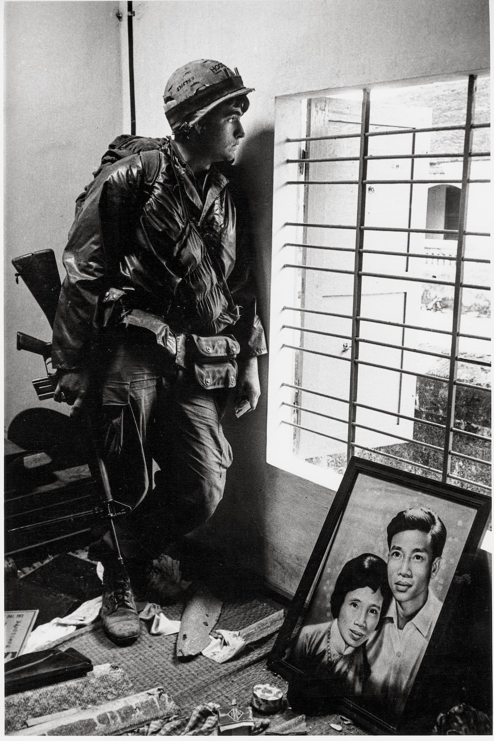 A soldier in uniform and helmet stands by a barred window, holding a rifle. On the floor beside him is a framed portrait of a man and woman, surrounded by scattered belongings.