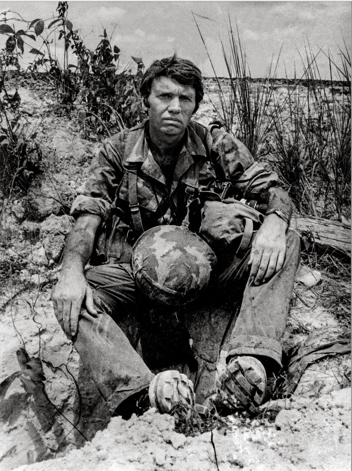 A tired soldier in a dirty uniform sits on the ground amid sand and sparse vegetation, holding his helmet in his lap and looking directly at the camera with a weary expression.