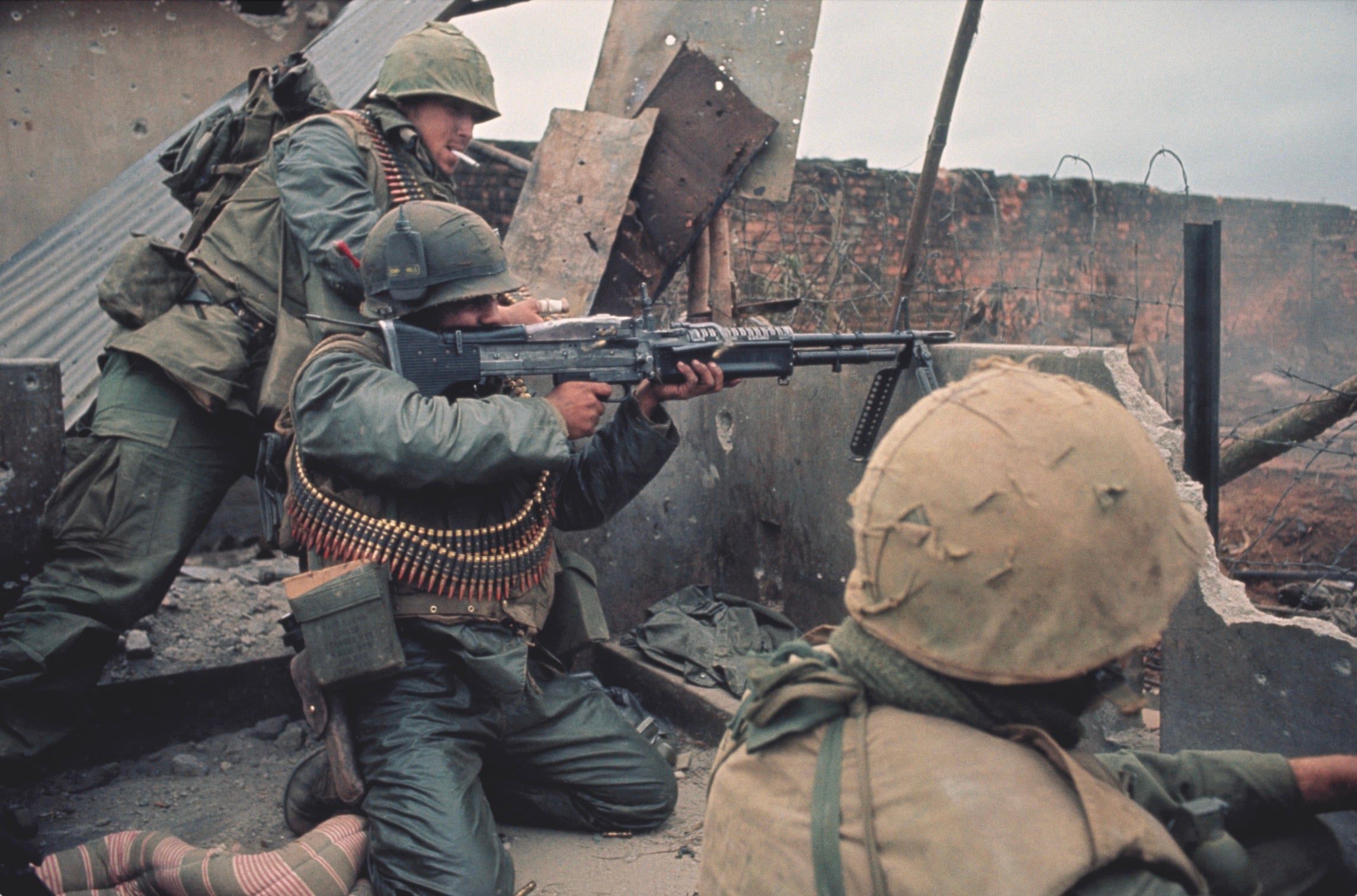Three soldiers in combat gear take cover behind rubble. One is aiming a machine gun, while another stands behind and the third crouches nearby. The scene appears tense and war-torn, with debris and ruined walls around them.