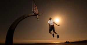 A basketball player in mid-air is about to dunk the ball on an outdoor court at sunset, with the sun glowing brightly behind him and the ocean visible in the background.