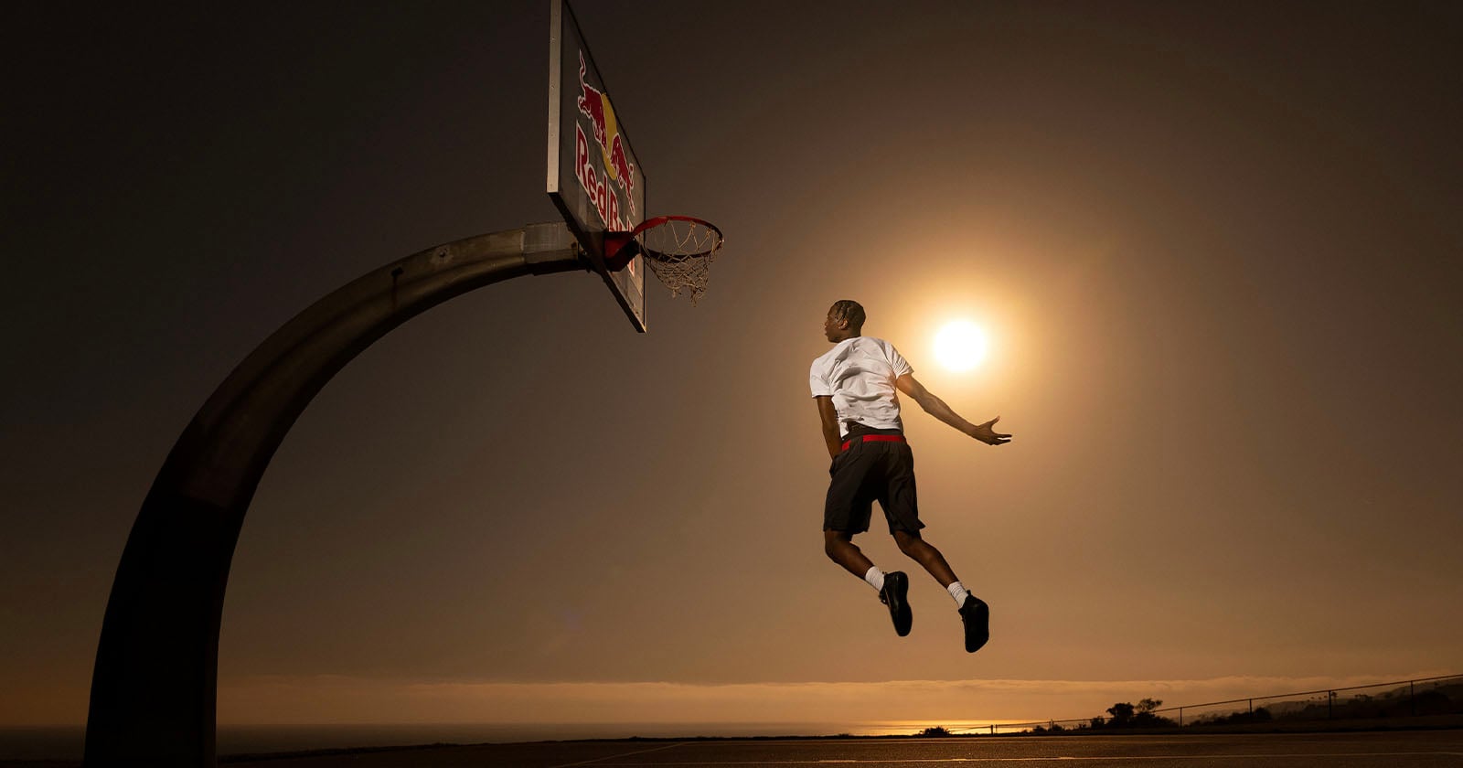 A basketball player in mid-air is about to dunk the ball on an outdoor court at sunset, with the sun glowing brightly behind him and the ocean visible in the background.