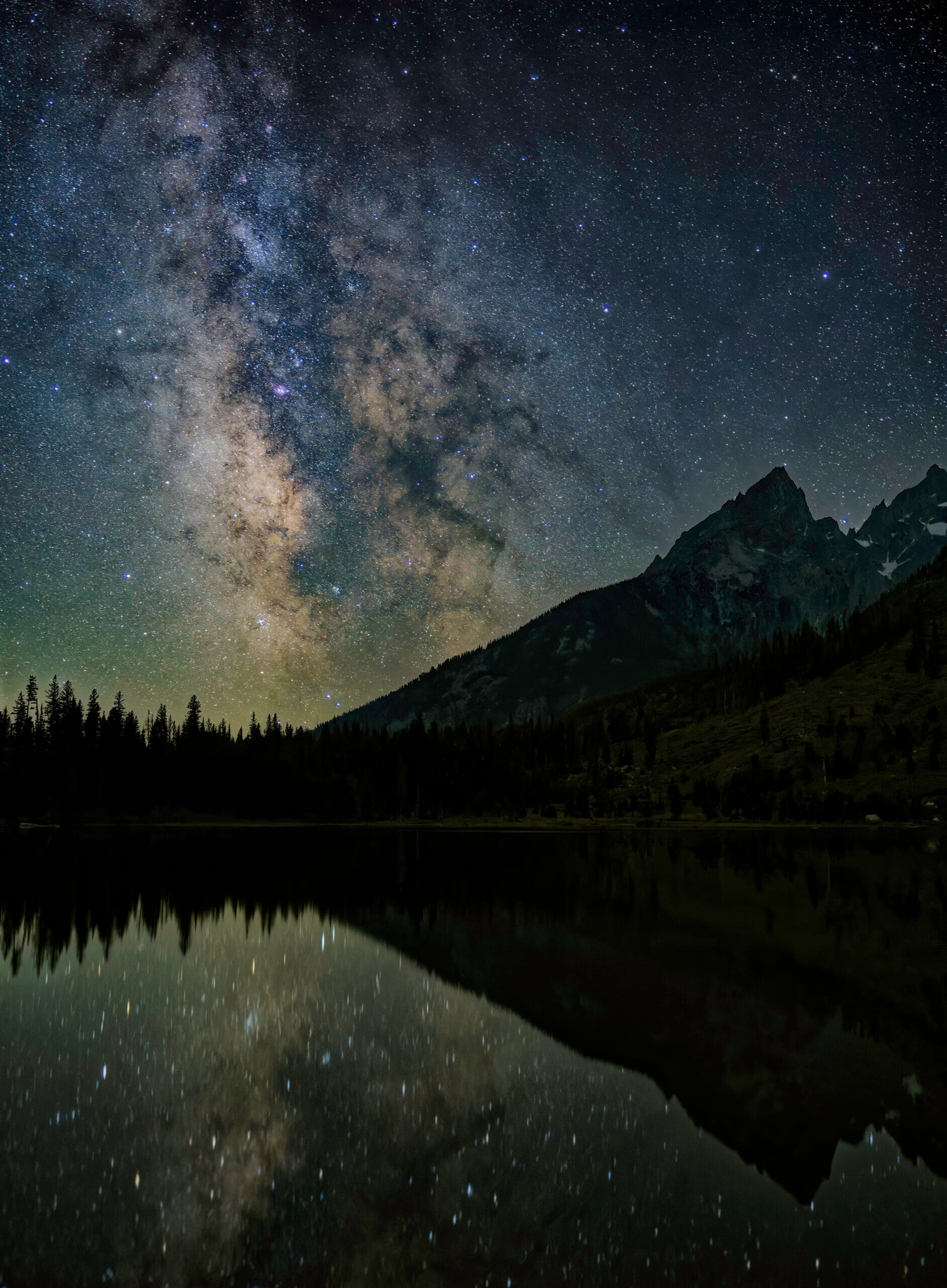 The Milky Way galaxy stretches across a star-filled night sky above a mountain range, with the scene and stars clearly reflected in a calm lake below.