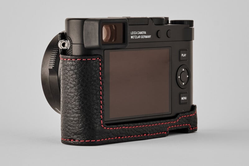 A black Leica camera with a textured black leather case and red stitching, shown from the back with its screen, buttons, and viewfinder visible against a plain gray background.
