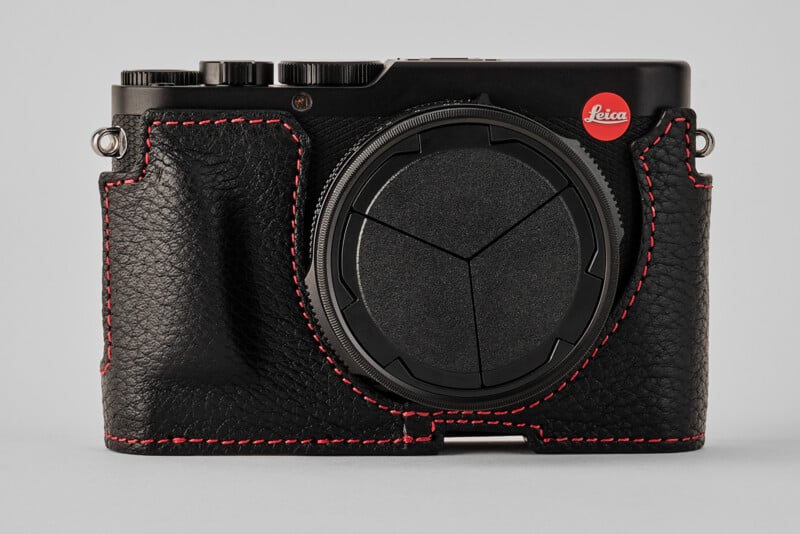 A black Leica camera with a textured leather case featuring red stitching, viewed from the front against a plain gray background.