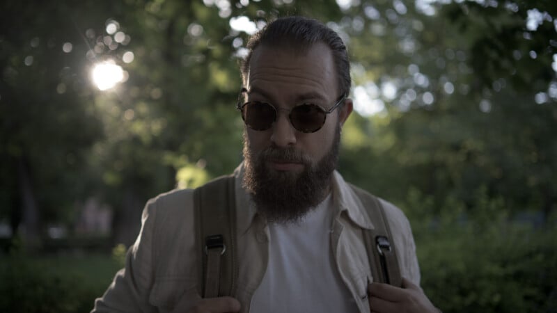 A bearded man wearing sunglasses and a light jacket stands outdoors, holding backpack straps, with sunlight and green foliage in the background.
