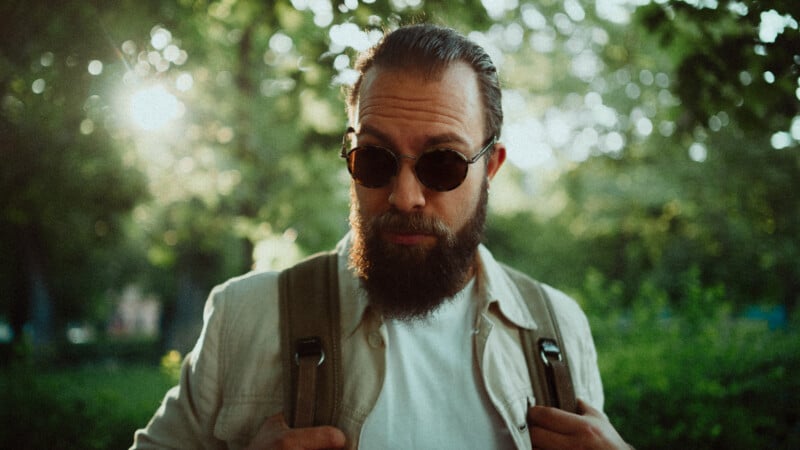 A bearded man wearing sunglasses and a light jacket stands outdoors, holding backpack straps, with sunlight and green foliage in the background.