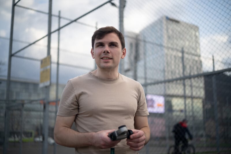 A man in a beige t-shirt stands outside holding a camera, with a chain-link fence and modern buildings in the background. A cyclist passes by in the distance. The scene appears to be in an urban environment.