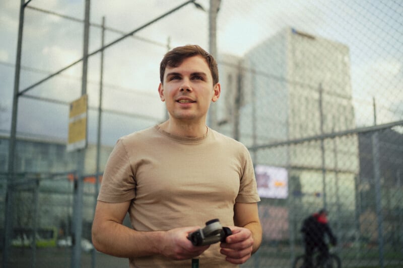 A young man in a beige t-shirt holds a game controller while standing on an outdoor basketball court with metal fencing. Modern glass buildings and a cyclist are visible in the background.