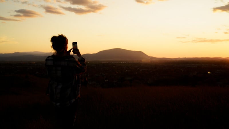A person stands in a field at sunset, taking a photo with a phone. The sky is orange and yellow, with a few clouds, and a mountain is visible in the distance.