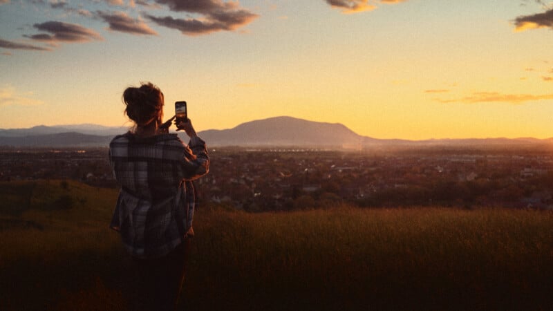 A person standing in a grassy field takes a photo of a scenic sunset over a city with mountains in the background, the sky glowing with orange and yellow hues.