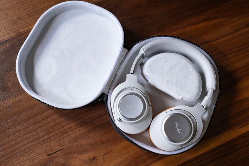 A pair of white over-ear headphones with silver accents rests in an open, white zippered carrying case on a wooden surface.