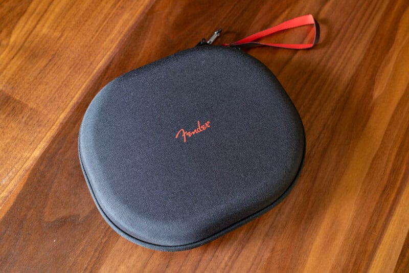 A black, oval-shaped zippered carrying case with a red "Fender" logo and a red strap is placed on a wooden surface.