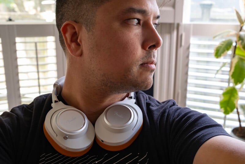 A person wearing a dark shirt looks to the side with a pair of white headphones resting around their neck. They are indoors, near a window with blinds and a potted plant in the background.