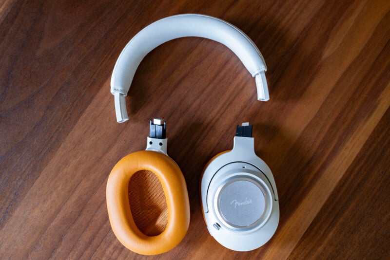 A pair of white and orange headphones with the ear cups detached, lying on a wooden surface. The brand name “Fender” is visible on one of the ear cups.