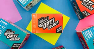 A box of Opti Colour 35mm film sits on a yellow paper, surrounded by other colorful Opti Colour film boxes on a blue background. The central box shows icons for weather conditions and "36 EXP ISO 200.