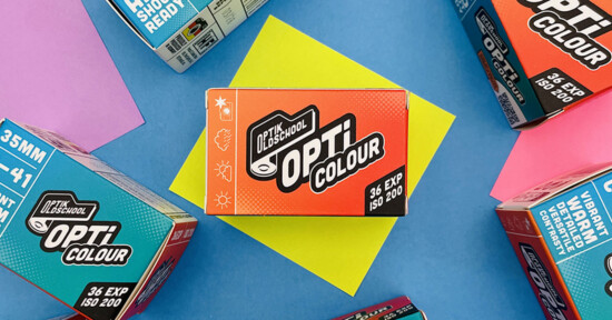A box of Opti Colour 35mm film sits on a yellow paper, surrounded by other colorful Opti Colour film boxes on a blue background. The central box shows icons for weather conditions and "36 EXP ISO 200.