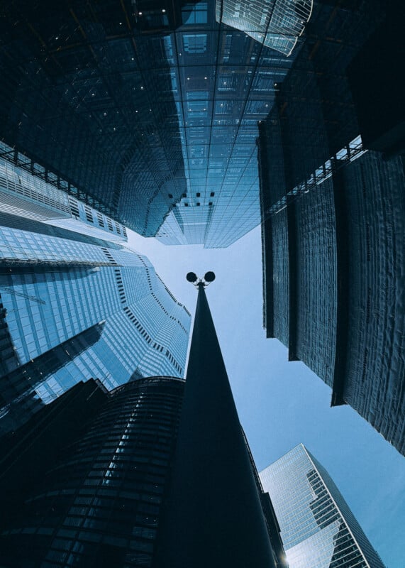 A view looking up at tall modern skyscrapers with reflective glass windows, converging towards the blue sky. A streetlamp stands in the center, surrounded by the towering buildings.