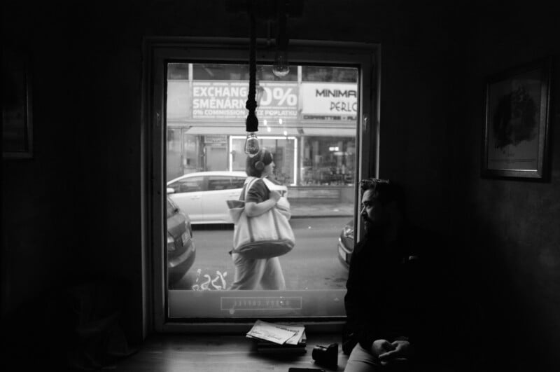 A person sits in a dimly lit room by a window, looking outside. Through the window, a person carrying bags walks past parked cars on a city street with shop signs visible in the background.