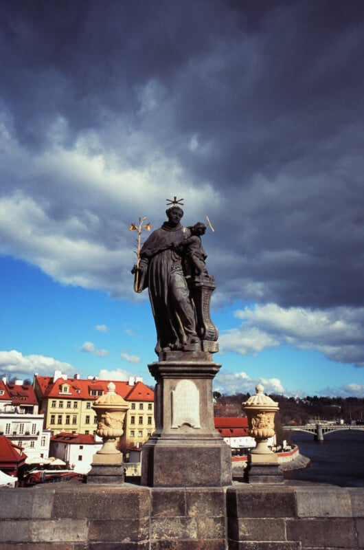 A statue of a saint holding a crucifix stands on a stone pedestal on a bridge, with ornate vases on each side. Colorful buildings and a river are visible in the background under a dramatic, cloudy sky.