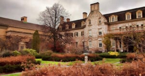 A large historic stone building with tall windows and dormers stands behind landscaped gardens with brown shrubs, winding paths, and a sundial under cloudy skies.