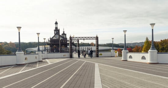 Wide pedestrian walkway leading to a decorative gate and pavilion, with several people walking toward it. Street lamps line the path, and trees with autumn foliage are visible in the background.