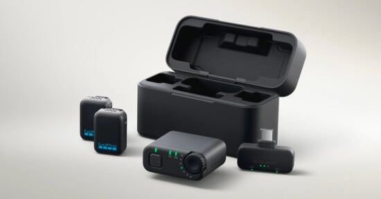 A GoPro wireless microphone system with two microphones, a receiver, a charging case, and a USB-C adapter, all shown on a light background. Green indicator lights are visible on the receiver and microphones.