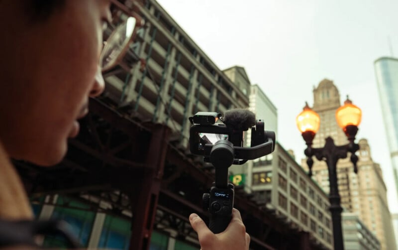 A person holds a camera mounted on a stabilizer, filming city buildings and a lit street lamp. The backdrop features elevated train tracks and tall skyscrapers under a cloudy sky.