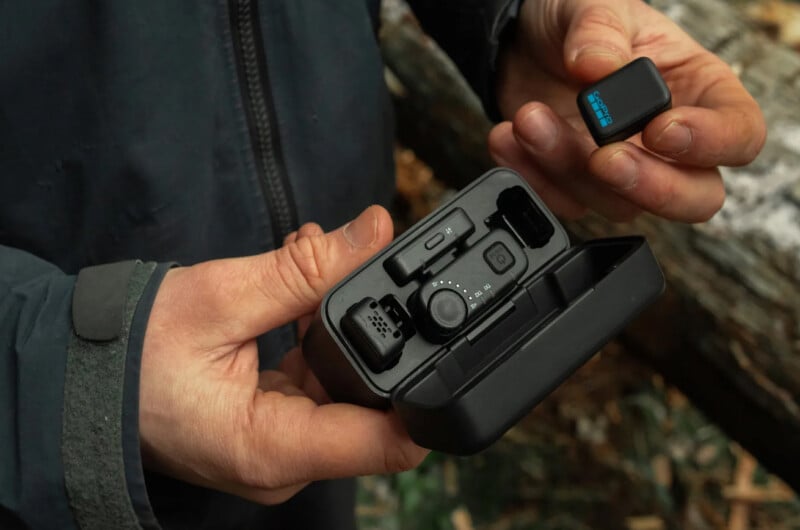 A person holds an open black case containing GoPro camera accessories in one hand and a small GoPro battery in the other, with a blurred outdoor background of leaves and a log.