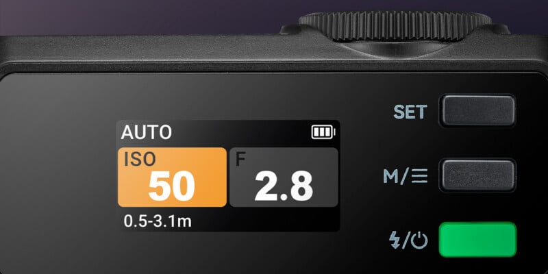 Close-up of a digital camera display showing settings: ISO 50, aperture f/2.8, Auto mode, battery icon, and distance range 0.5-3.1m. Buttons labeled SET, M/, and a green power button are visible.