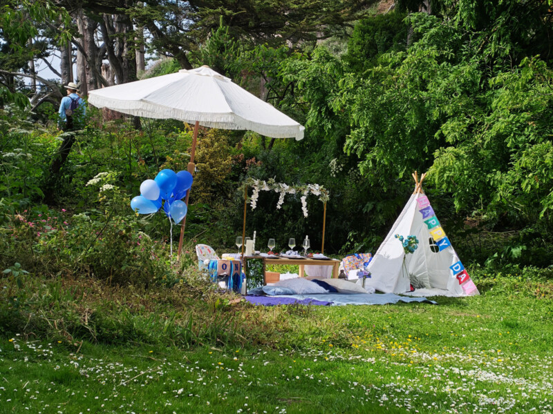 A picnic setup in a grassy area with a white umbrella, blue balloons, a low table with place settings, cushions, and a decorated white teepee surrounded by trees and greenery. A person stands in the background among the trees.