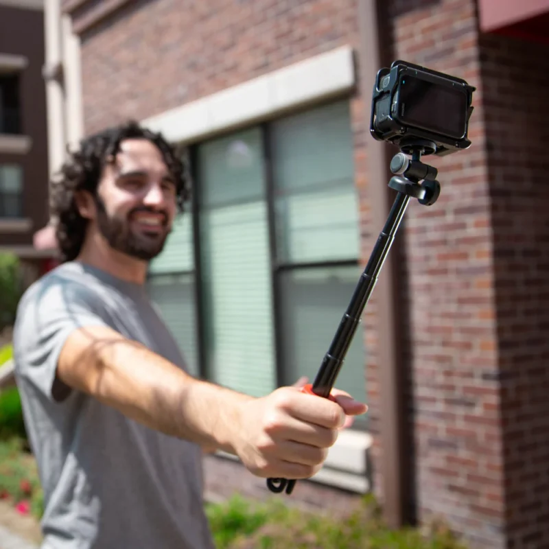 A person with curly hair and a beard is smiling while holding a camera mounted on a selfie stick outdoors in front of a brick building.