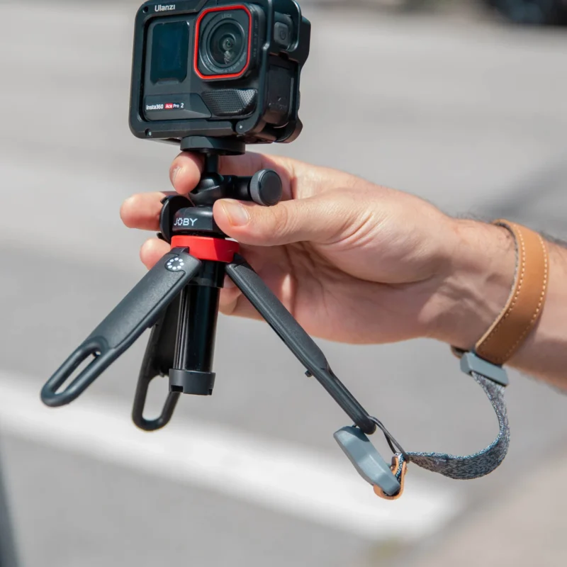 A person holds a small tripod-mounted action camera, with the camera facing forward. The tripod has black legs, a red ring, and a wrist strap. The background is outdoors and slightly blurred.