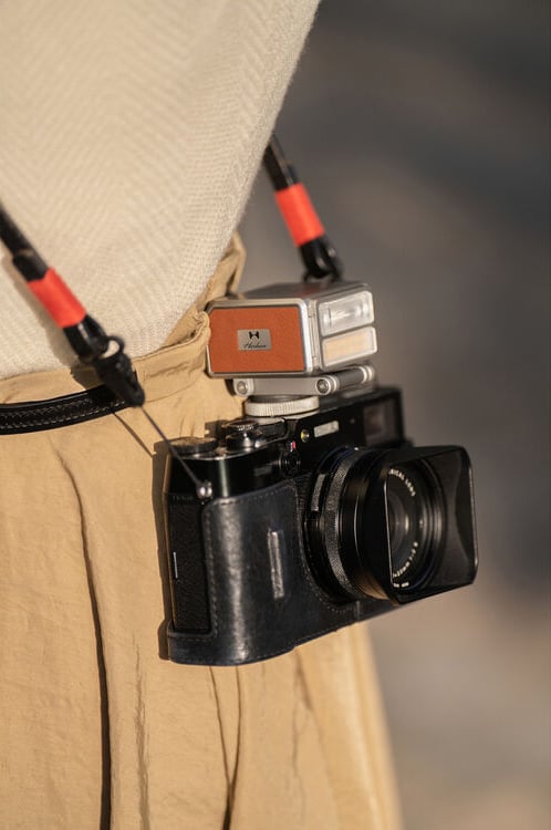 A close-up of a person wearing a beige outfit with a black camera and a small flash attached, both hanging from a black and red strap around their neck.