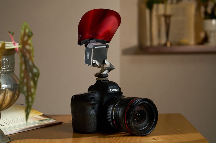 A Canon DSLR camera with a large lens sits on a wooden table. A red flash diffuser is attached to the camera’s external flash. In the background, blurred books and a plant are visible.